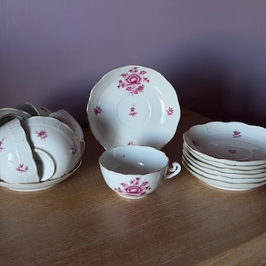 May include: A collection of white porcelain teacups, saucers, and plates with a pink floral design. The teacups have a delicate handle and the plates have a scalloped edge. The set is arranged on a wooden surface.