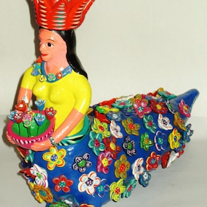 May include: A colorful ceramic figurine of a woman wearing a red crown and holding a basket of flowers. The woman is sitting on a blue base decorated with colorful flowers.