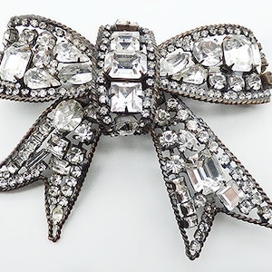 May include: A silver-toned bow brooch with a large, clear rhinestone center and smaller rhinestones throughout. The bow is made of metal and has a textured finish.
