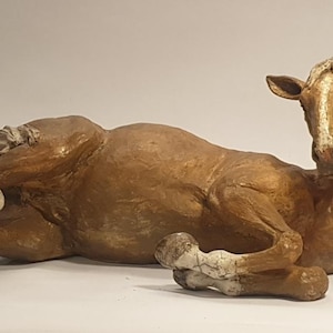 May include: A ceramic sculpture of a brown horse lying down with its head turned to the side. The horse has white markings on its legs and face.