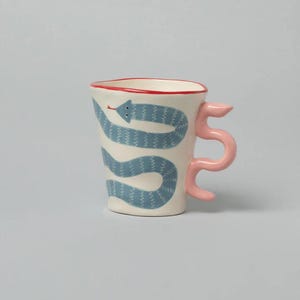 May include: A ceramic mug with a white base, featuring a blue snake design wrapping around the body. The rim is red, and the handle is a pink, stylized '5'. The mug is a unique and artistic piece.