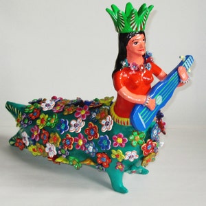 May include: A colorful ceramic figurine of a woman playing a blue ukulele. The figure has a red top, a green skirt, and a green crown. The lower body is a teal shape covered in colorful flowers. The figurine is a unique decorative item.