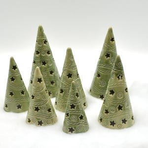 May include: A collection of seven green ceramic Christmas trees with star-shaped cutouts. The trees have a conical shape and are decorated with etched spiral and star patterns. The trees are arranged on a white surface.