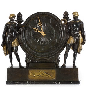 May include: An antique bronze clock with a dark finish, featuring two sculpted figures flanking a circular clock face. The clock has gold accents and a rectangular base with a relief of a reclining figure. The clock hands point to 10:08.