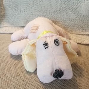 May include: A handmade plush dog toy in soft pink fabric. The dog has floppy ears, black embroidered eyes, and a black nose. A yellow ribbon is around its neck, and a small patch is sewn on its back.