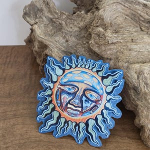 Op de afbeelding: Een gestanste sticker met een gestileerde zon met een blauw en oranje gezicht. De zon heeft golvende, vlamachtige stralen die naar buiten uitsteken in tinten blauw en lichtgroen. Het centrale gezicht bevat gedetailleerde illustraties van ogen.