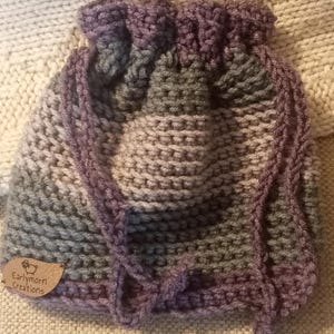 Könnte beinhalten: Eine lila, graue und blaue gehäkelte Babyhaube mit einem braunen Etikett mit der Aufschrift "Earlymorn Creations".