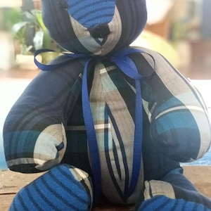 May include: A blue and gray plaid teddy bear with a blue ribbon around its neck. The bear is sitting on a wooden surface.