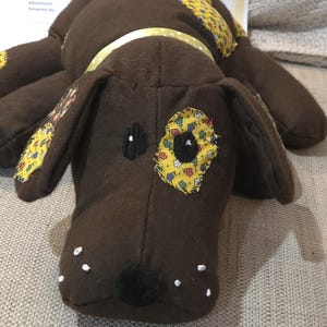 May include: A brown stuffed dog with yellow floral patches and a yellow collar. The dog has black embroidered eyes and a black nose. White dots are on the snout. The dog's birthday is April 23, 2026.