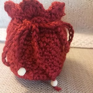 Può includere: Un sacchetto con coulisse fatto a mano, all'uncinetto rosso. Il sacchetto è rotondo con una parte superiore a balze e una chiusura con coulisse. Due perline bianche sono attaccate al fondo del sacchetto. Il sacchetto è realizzato con un filato spesso e strutturato.