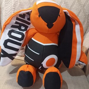 Könnte beinhalten: Orange-schwarzes Plüschkaninchen mit weißen Akzenten. Die Ohren tragen den Schriftzug "Giroux". Der Körper hat ein schwarz-weißes Design. Das Kaninchen sitzt auf einer beigen Oberfläche.