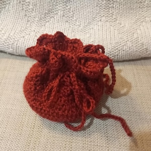 May include: A small, round, crocheted pouch in a deep red hue. The pouch is gathered at the top with a drawstring closure. The texture of the crochet is visible, and the pouch appears to be handmade. The background is a neutral, textured fabric.