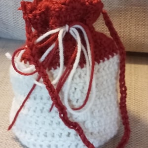 May include: A handmade crochet drawstring bag in red and white. The top and drawstring are red, while the body is white. The bag has a braided red strap. The bag is made of yarn.