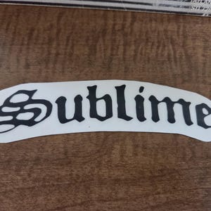 Puede incluir: Una calcomanía de vinilo negra con la palabra Sublime escrita en una fuente gótica estilizada. Las letras están sobre un fondo blanco con una forma curva y arqueada que sigue el contorno del texto, creando un diseño de pegatina distintivo.