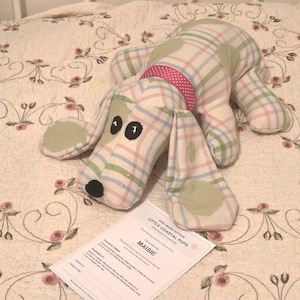 May include: A stuffed dog toy made from plaid fabric in shades of pink, blue, and green, featuring a pink polka-dot collar, black embroidered eyes, and a black nose. A white adoption certificate paper labeled Little Coastal Pups is included.