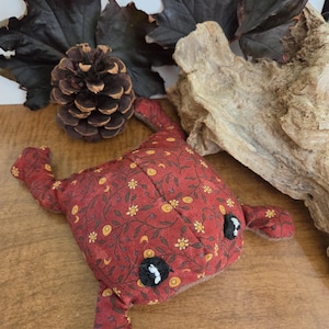 Könnte beinhalten: Ein Stofffrosch aus rotem Stoff mit einem sich wiederholenden Muster aus gelben Blumen, Ranken und Mondsicheln. Der Frosch hat zwei schwarze, kreisförmige Augen mit weißen Stickakzenten, vier Gliedmaßen und einen abgerundeten Körper aus Stoff.