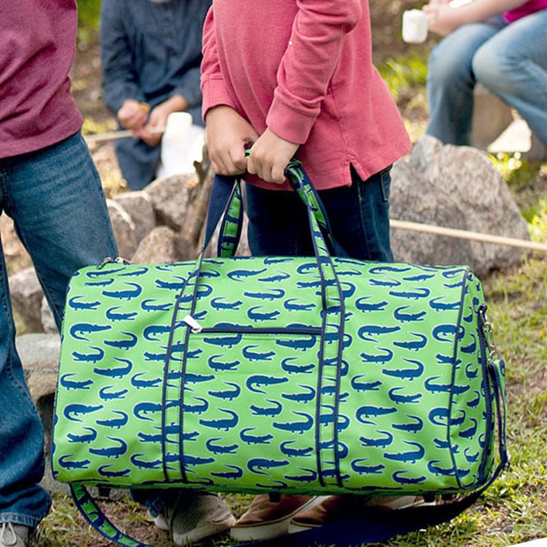 Pu&ograve; includere: Una borsa da viaggio verde con un motivo di alligatori blu. La borsa ha una tasca frontale con cerniera, finiture blu navy e manici. La borsa &egrave; tenuta da una persona.