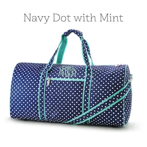 May include: Navy blue duffel bag with white polka dots and mint green accents. The bag features a zipper closure, a shoulder strap, and handles. The text "Navy Dot with Mint" is displayed at the top of the image. The bag has an embroidered monogram.