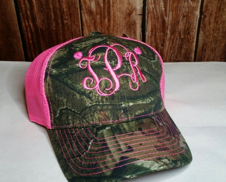 Może przedstawiać: Czapka trucker w kolorze r&oacute;żowym i zielonym kamuflażu z haftowanym r&oacute;żowym monogramem "JPR" z przodu.
