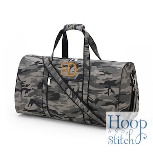 Pu&ograve; includere: Borsa da viaggio con motivo mimetico grigio e nero, con tracolla e manici superiori. La borsa ha uno scomparto principale con cerniera e una tasca esterna con un monogramma arancione.