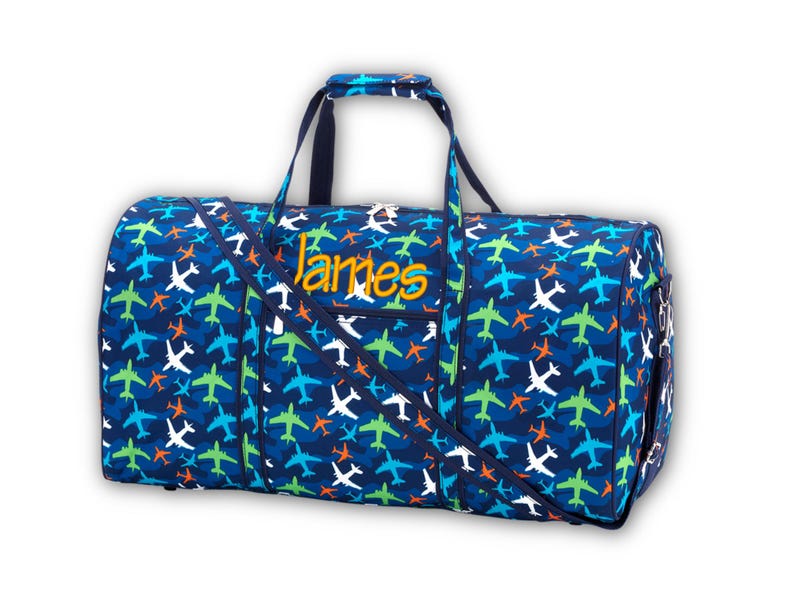 Pu&ograve; includere: Una borsa da viaggio blu con un motivo ripetuto di aerei bianchi, verdi, blu e arancioni. La borsa ha una tracolla e manici. Il nome "James" &egrave; ricamato in giallo sul davanti.