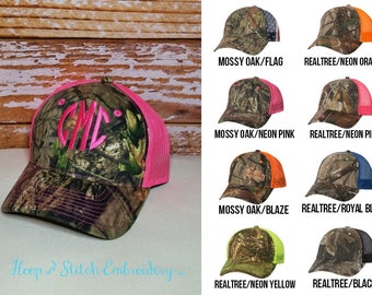 May include: A selection of camouflage trucker hats with different colored mesh back panels. The hats are available in Mossy Oak and Realtree patterns with colors such as neon pink, neon orange, blaze orange, royal blue, neon yellow, and black. The text "Hoop and Stitch Embroidery" is visible at the bottom of the image.