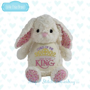 May include: A plush white bunny with pink floral patterned ears and feet. The bunny has black eyes and a pink nose. Embroidered on the bunny's chest is the phrase "CHILD OF THE ONE TRUE KING" in gold and pink.