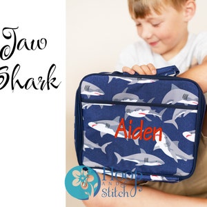 May include: A blue lunch bag with a shark print and the name "Aiden" embroidered in red. The bag has a zip closure and a top handle.