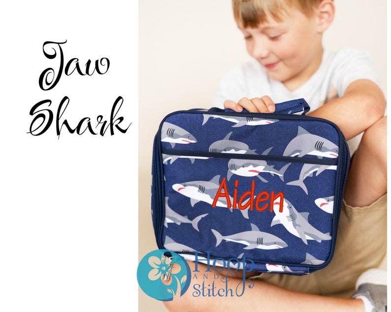 May include: A blue lunch bag with a shark print and the name "Aiden" embroidered in red. The bag has a zip closure and a top handle.