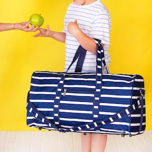 Pu&ograve; includere: Una borsa da viaggio a righe blu navy e bianche con tracolla e tasca con cerniera. La borsa &egrave; tenuta da una persona che indossa una maglietta a righe bianche e blu. Una mela verde viene offerta alla persona.