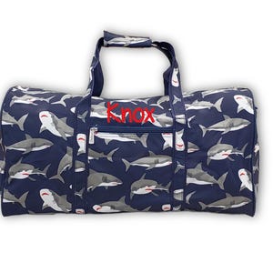 Pu&ograve; includere: Borsa da viaggio blu navy con un motivo di squali grigi e bianchi. La borsa ha il nome "Knox" ricamato in rosso e una tasca con cerniera. Include due manici e una tracolla.