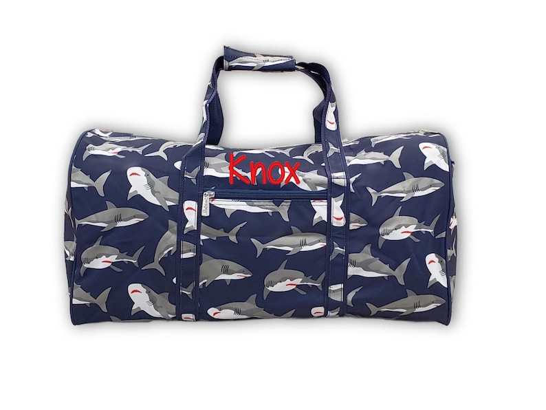 Pu&ograve; includere: Borsa da viaggio blu navy con un motivo di squali grigi e bianchi. La borsa ha il nome "Knox" ricamato in rosso e una tasca con cerniera. Include due manici e una tracolla.