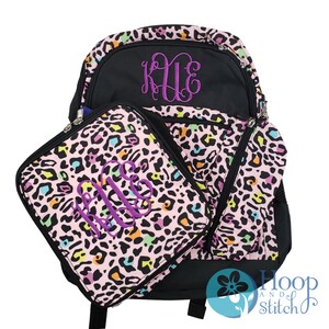 May include: A pink and black leopard print backpack with a matching lunch bag. Both have a purple monogram "KUE" embroidered on them.