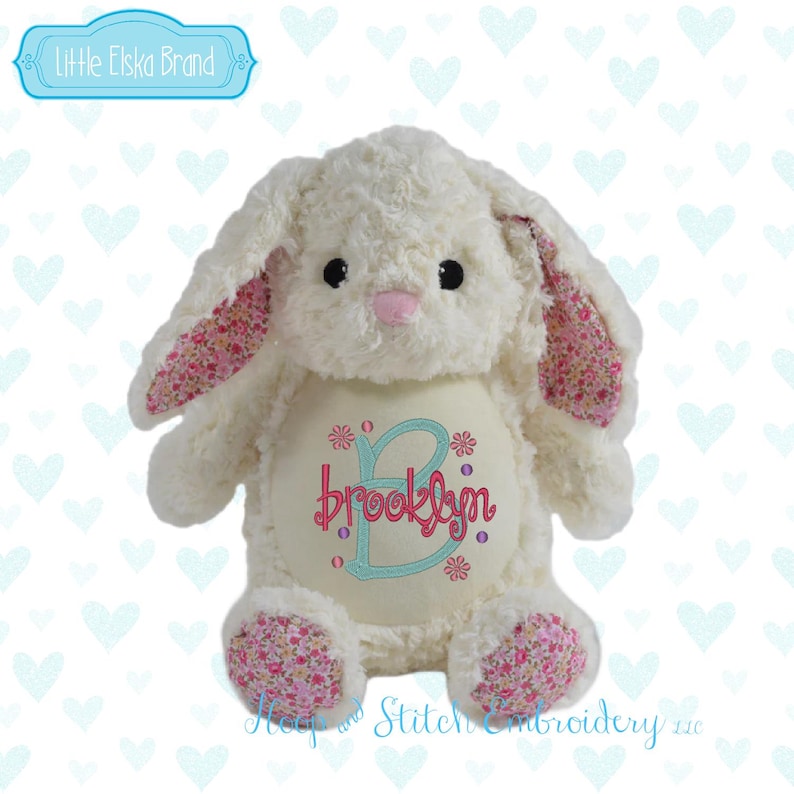 May include: A plush white bunny rabbit with long ears and a pink nose. The inner ears and foot pads feature a floral pattern. The bunny has the name "Brooklyn" embroidered on its belly in pink and teal, with floral accents. The brand name "Little Elska Brand" is in the upper left corner.