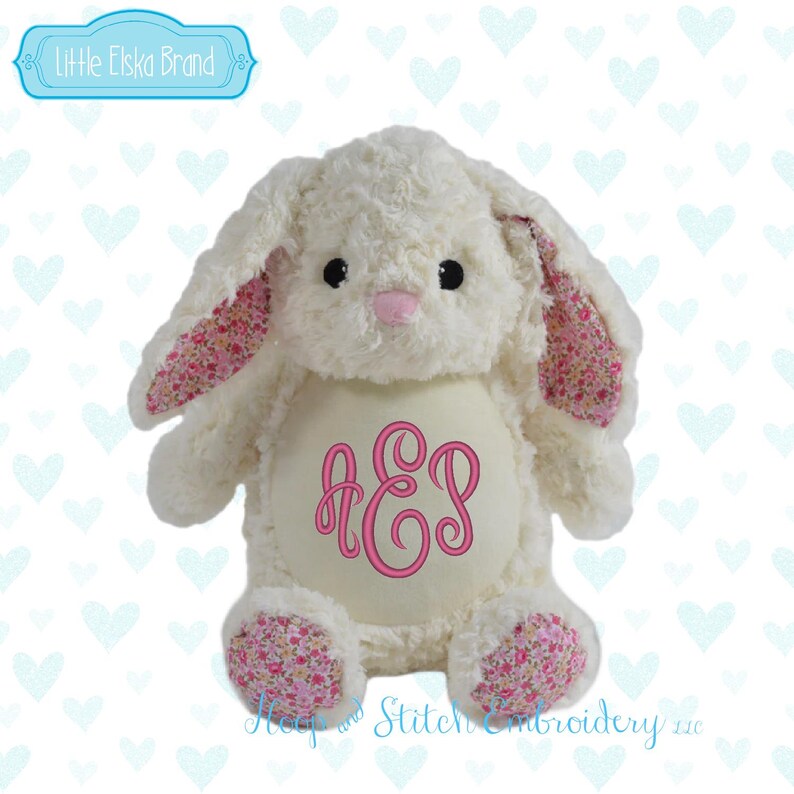 May include: A plush white bunny rabbit with pink floral patterned ears and feet. The bunny has a pink nose and black eyes. The belly features a pink monogram. The brand name "Little Elska Brand" is in a blue banner.