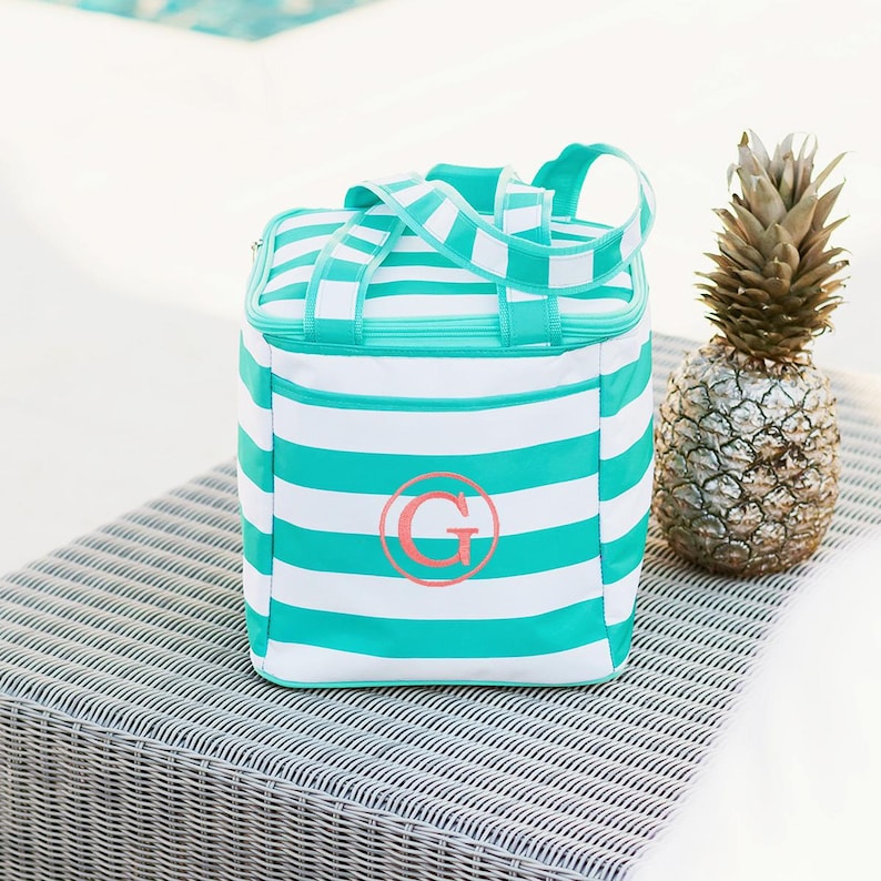 May include: A white and teal striped cooler bag with a teal handle and a pink monogrammed "G". The bag is sitting on a gray woven surface next to a gold-painted pineapple.