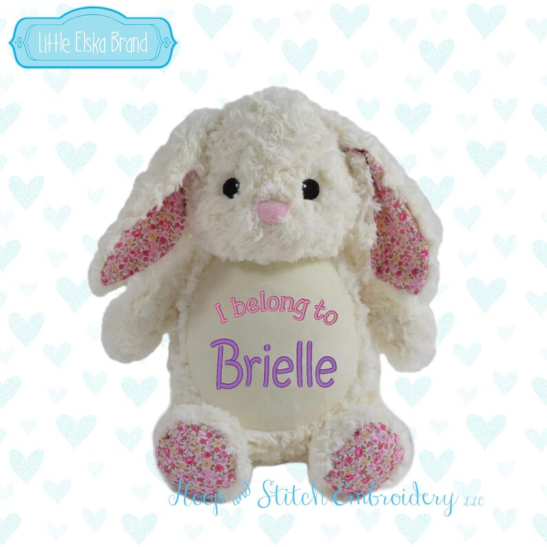 May include: A plush white bunny with floppy ears and floral patterned accents on the inner ears and feet. The bunny has embroidered text that reads "I belong to Brielle". The bunny is a stuffed animal.