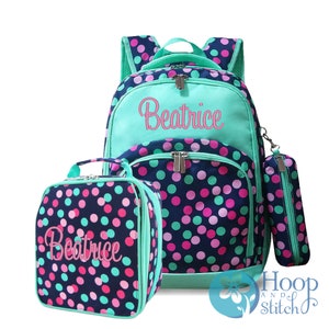 May include: A turquoise and navy blue polka dot backpack set with a matching lunch bag and pencil case. The backpack has a large zippered pocket on the front with the name "Beatrice" embroidered in pink.