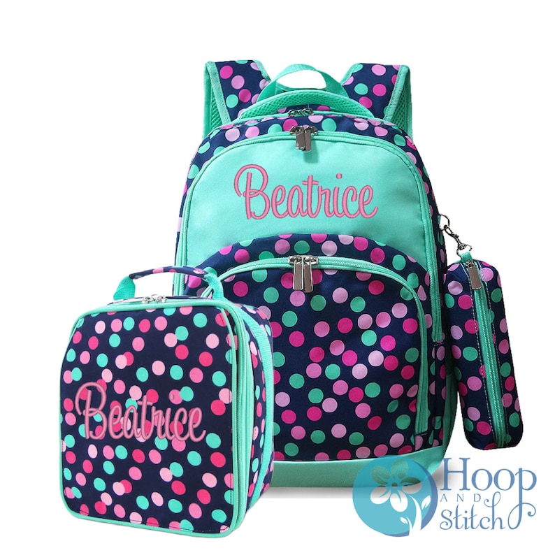 May include: A turquoise and navy blue polka dot backpack set with a matching lunch bag and pencil case. The backpack has a large zippered pocket on the front with the name "Beatrice" embroidered in pink.