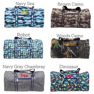 May include: Six duffel bags with various patterns and text. Designs include blue and white brushstrokes, brown camouflage, robot graphics, woodland camouflage, gray chambray, and dinosaur illustrations. Each bag has a different name embroidered on it.