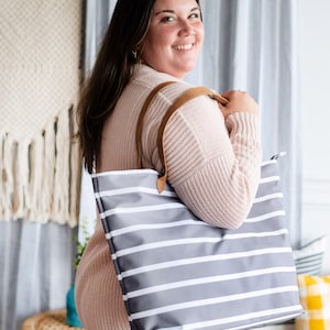 May include: A gray tote bag with white horizontal stripes and brown leather handles. The bag is held by a person wearing a light pink sweater. The background includes a woven wall hanging and striped curtains.