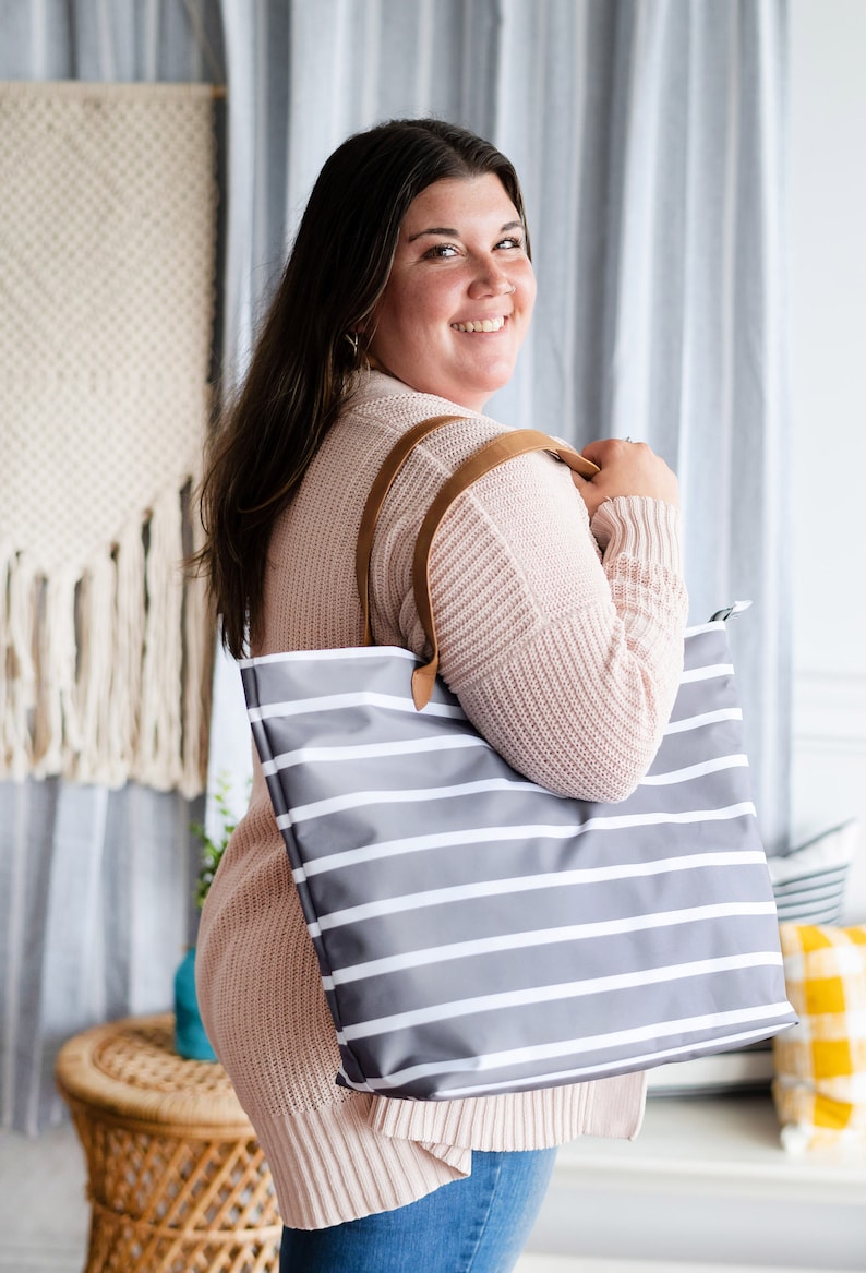 May include: A gray tote bag with white horizontal stripes and brown leather handles. The bag is held by a person wearing a light pink sweater. The background includes a woven wall hanging and striped curtains.