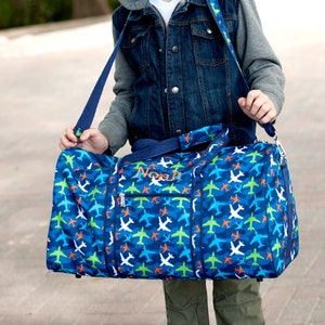 Pu&ograve; includere: Una borsa da viaggio blu con un motivo di aerei bianchi, verdi e arancioni. La borsa ha una tracolla blu navy e il nome "Noah" ricamato in verde chiaro. La borsa &egrave; tenuta da una persona.