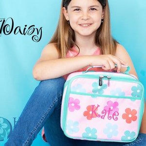 May include: A pink lunch bag with a floral pattern and the name "Kate" embroidered on the front. The bag has a green zipper and a green handle.