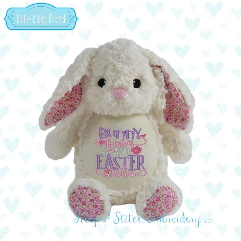 May include: A plush white bunny with pink floral patterned ears and paws. The bunny has black eyes and a pink nose. The text "Bunny kisses and Easter wishes" is embroidered on the bunny's chest. The brand name "Little Elska Brand" is in the top left corner.