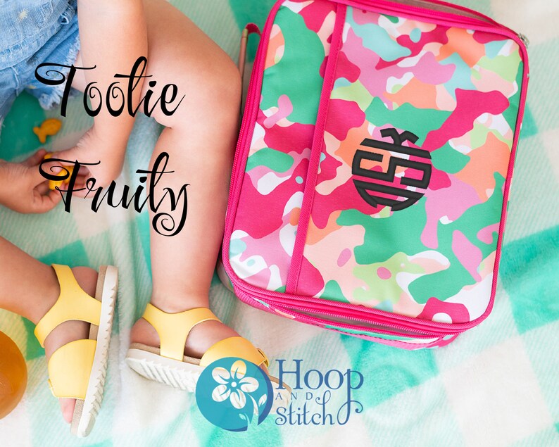May include: A pink and green camouflage lunch bag with a black monogram. The bag is on a turquoise and white blanket. The text "Tootie Fruity" is written in black cursive on the left side of the image. The text "Hoop and Stitch" is written in black cursive on the right side of the image.