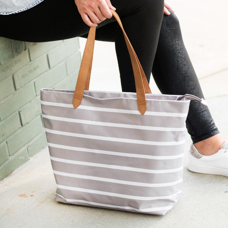May include: A gray and white striped tote bag with brown leather handles. The bag is held by a person wearing black leggings and white sneakers. The tote bag is a light gray color with horizontal white stripes.