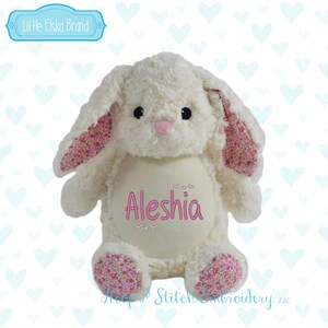 May include: A plush white bunny rabbit with pink floral patterned ears and feet. The bunny has black eyes and a pink nose. The name "Aleshia" is embroidered in pink on the bunny's belly. The brand name "Little Elska Brand" is in a blue banner.