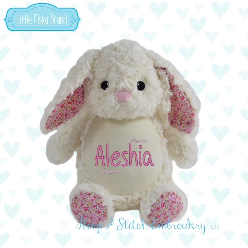 May include: A plush white bunny rabbit with pink floral patterned ears and feet. The bunny has black eyes and a pink nose. The name "Aleshia" is embroidered in pink on the bunny's belly. The brand name "Little Elska Brand" is in a blue banner.