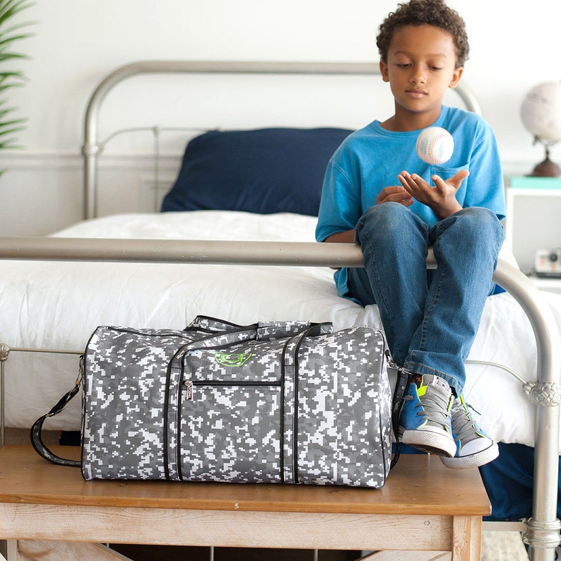 Pu&ograve; includere: Una borsa da viaggio mimetica digitale grigia e bianca con finiture nere e un logo verde. La borsa ha una tracolla e si trova su una superficie di legno. Una persona &egrave; seduta su un letto sullo sfondo.